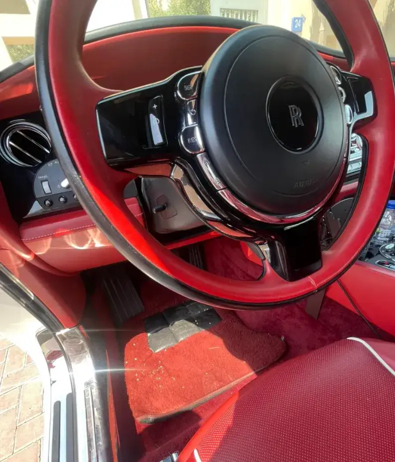 Close-up of a Rolls-Royce interior with a red-and-black steering wheel, red upholstery, and visible debris on the driver's floor mat.