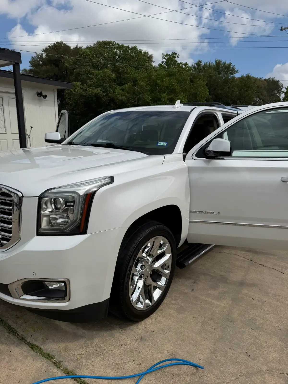 A white Denali SUV parked on a concrete driveway with both front doors open. Its front headlight and chrome wheel are prominent, and a blue hose lies across the foreground.