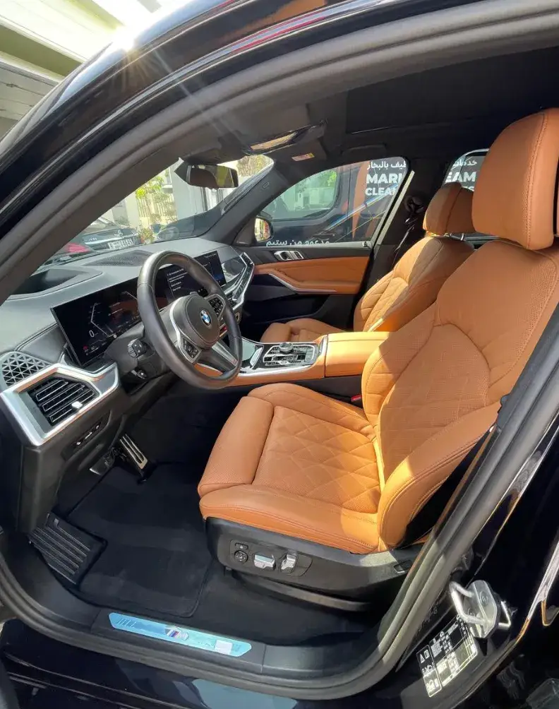 View through an open BMW driver's door showing tan quilted leather front seats, a black dashboard, steering wheel, center console and dark floor mats.