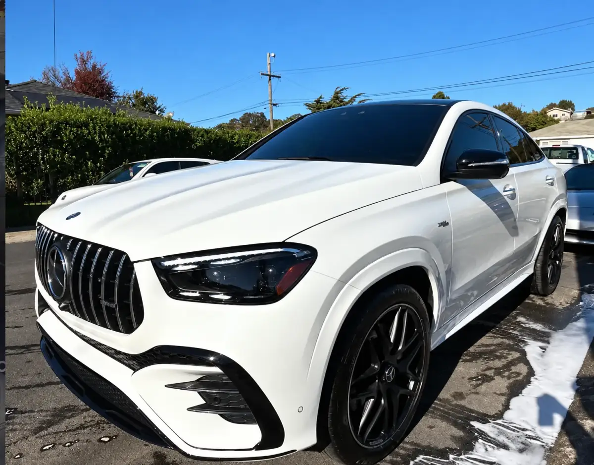 A white Mercedes-Benz SUV with black wheels parked outdoors on wet pavement with soap suds around it.